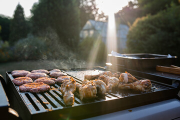 Close up of a bbq grill with meats cooking on them, sun flare in the background is catching the smoke