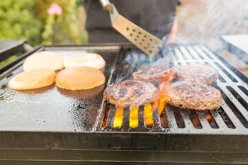 A happy young man cooking meat burgers on barbecue grill - leisure, food, people and holidays concept