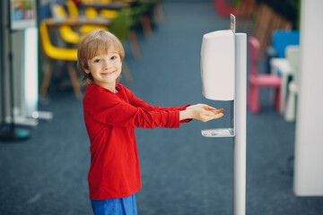 Child smiling boy kid using automatic alcohol gel dispenser spraying on hands sanitizer machine...