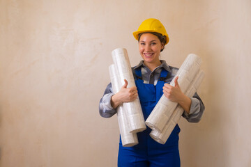 A girl in a blue construction worker's jumpsuit and a yellow hard hat holds wallpaper in her hands, smiles and gives a thumbs-up against a gray stucco wall