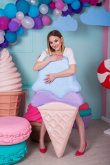 Portrait of young woman in pink dress holding big ice cream and posing on decorated background. Amazing sweet-tooth girl surrounded by toy sweets.