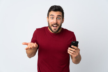 Young handsome man with beard using mobile phone isolated on white background with shocked facial expression