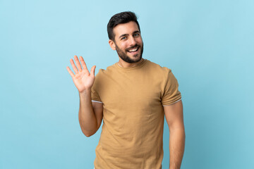 Young handsome man with beard over isolated background saluting with hand with happy expression