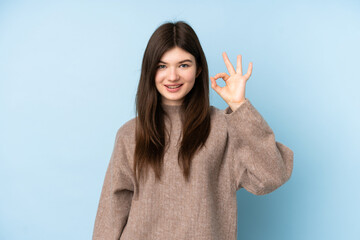 Young Ukrainian teenager girl wearing a sweater over isolated blue background showing ok sign with fingers