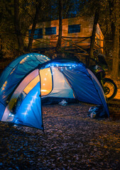 Big blue tent, glows. garlands and lighting in the camp. Camping. Motorcycle and hammock. Parking in the woods. Travel and vacation. Equipment for tourism. vertical photo. Marmore, Italy