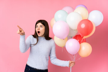 Young Ukrainian teenager girl holding lots of balloons over isolated pink background pointing away