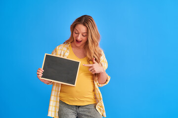 attractive, pregnant woman holds board with inscriptions in her hands. happy mom-to-be holds blank advertising banner and points at it with her finger while standing on blue background, copy space
