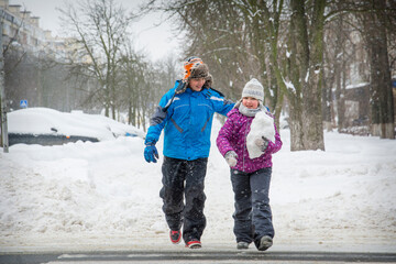 In winter, on the street in snowy weather, a boy and a girl cross the road along a pedestrian crossing.