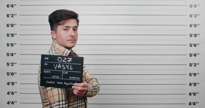 Side profile mugshot of male arrested looking to camera while standing in front of police metric lineup wall. Young guy turning head while holding sign for photo. Concept of crime.