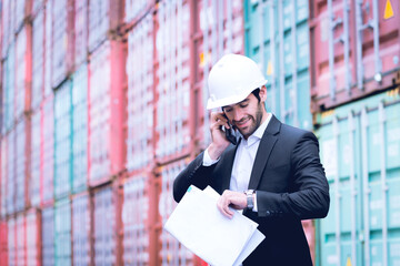 Handsome business man wears hardhat and holding blueprint and looking at his watch while talking about plan via cell phone with cargo container stacked in background. Concept of working at site.