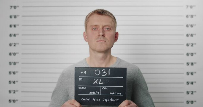 Portrait of adult man holding cardboard while being photographed in police department. Male person posing, raising head and looking to camera. Concept of mugshot and crime.