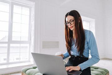 Attractive pretty young woman in eyeglasses freelancer using laptop computer at home, typing text and drinking coffee.  