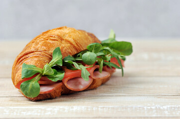 Croissant sandwich.  Sandwich filling with ham, tomato slices, arugula leaves and corn salad.  Light background and wooden table surface.  Close-up.  Free space for text placement.