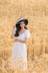 Naklejka na ściany i meble Happy young beautiful Asian woman in white dress and straw hat standing in the middle of the barley field on a bright sunny evening