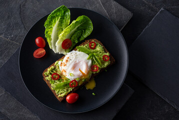 Fresh spring sandwich on a sliced gluten free sunflower bread. Red and green vegetables on black slate. Poached egg, avocado, chili and spring onion garnished with plum tomatoes