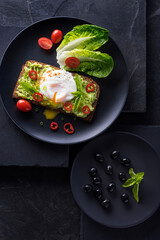 Fresh spring sandwich on a sliced gluten free sunflower bread. Red and green vegetables on black slate. Poached egg, avocado, chili and spring onion garnished with plum tomatoes