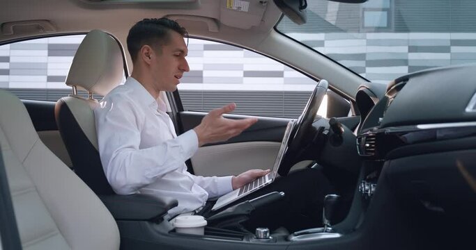 Portrait of young businessman man talking by videoconference using laptop while sitting in the car. Male entrepreneur greets by video chat, remote work concept.
