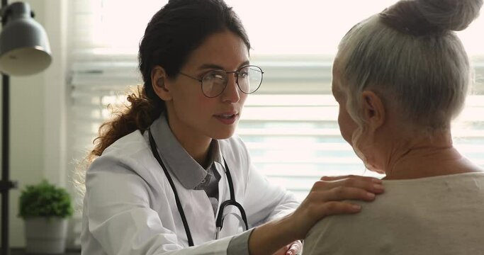 Confident young female general practitioner doctor in glasses stroking shoulder of elderly woman, supporting or giving professional psychological help at checkup meeting in hospital. medicare concept.