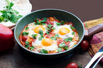Traditional dish of israeli cuisine Shakshuka. Fried eggs with tomatoes, peppers, onions, cilantro and pita in a pan on dark brown background