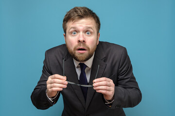 Shocked, frightened business man in a suit has taken off glasses and looks with big eyes opening mouth. Blue background.