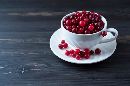 White Cup Full Of Rape Red Cranerry On The Saucer With Berries Too On The Dark Wooden Background. Healthy Nutrition. Copy Space