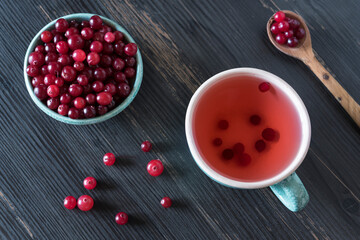 Fresh cranberry in a small ceramic bowl, cup with berry tea, wooden spoon and a few scattered berries on the dark grey wooden background. Top view. Flat lay