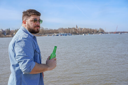 Bearded handsome young man standing on the river bank holding bottle of beer in his hand. Relaxing on his weekend vacation