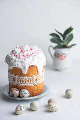  Traditional homemade Easter cake, decorated with glaze, pastry sprinkles and lace ribbon on a blue plate on a white table with quail eggs. Happy Easter. Front view.