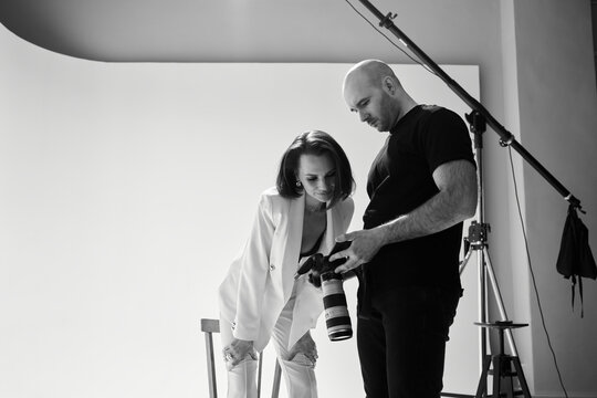 Fashion photography in a photo studio. Professional male photographer taking pictures of beautiful woman model on camera, black and white backstage