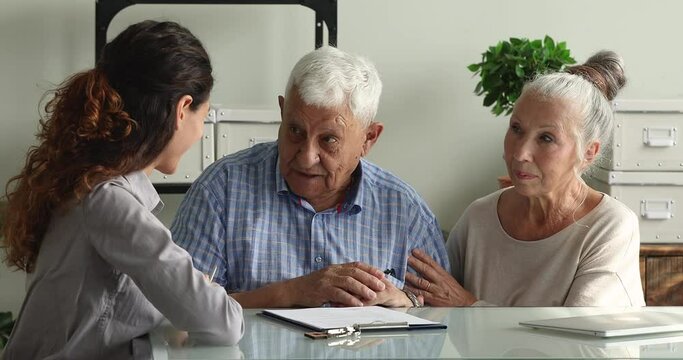 Professional young female real estate agent explaining house purchase contract details to older mature family couple, giving keys from own accommodation after signing agreement at office meeting.