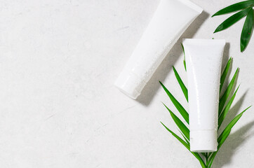 White unbranded cosmetic tubes with waterdrops with fresh palm leaves on white marble background. Organic skin care cosmetic concept.