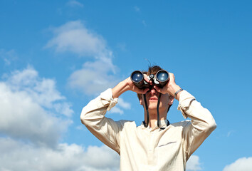 A young caucasian male looking at a view with binoculars under a sky with clouds