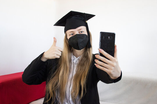 A happy female graduate in quarantine taking a selfie with her phone wearing a medical face mask and academic cap