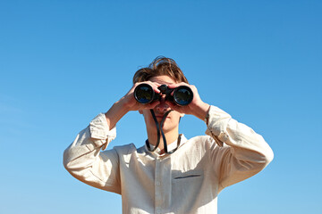 A young caucasian male looking at the camera with binoculars under the bright blue sky