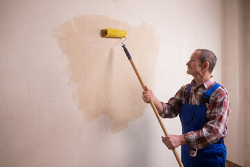An elderly man a painter with a mustache a construction worker in a blue uniform and a plaid shirt holds a tool in his hand a paint roller and smears glue on a plastered wall