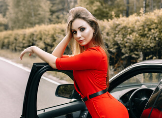 A beautiful young girl in a red overalls stands by a black car on an empty road in the forest