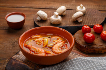 Tom yam soup with shrimps in a plate on the table on a napkin next to a bowl of coconut milk mushrooms and tomatoes.