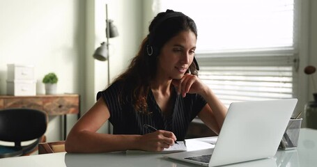 Smiling millennial woman in headphones learning foreign language distantly or studying on online courses, discussing educational material with teacher using computer video call app, taking notes. - Powered by Adobe