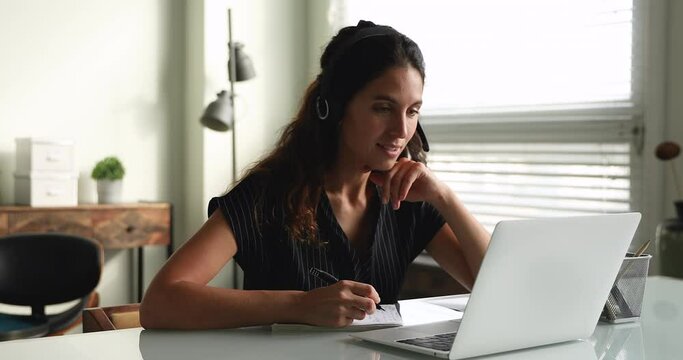 Smiling millennial woman in headphones learning foreign language distantly or studying on online courses, discussing educational material with teacher using computer video call app, taking notes.