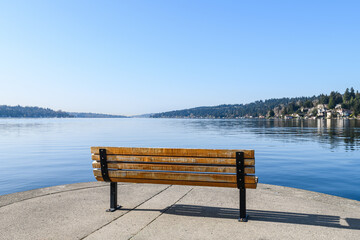 Naklejka na ściany i meble Bench at Lake Washington looks out onto smooth lake on a blue sky morning with no people to occupy the seat