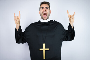 Young hispanic man wearing priest uniform standing over white background shouting with crazy...