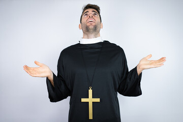 Young hispanic man wearing priest uniform standing over white background clueless and confused...