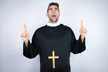Young hispanic man wearing priest uniform standing over white background amazed and surprised...