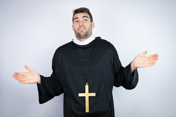 Young hispanic man wearing priest uniform standing over white background clueless and confused...