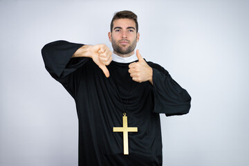 Young hispanic man wearing priest uniform standing over white background Doing thumbs up and down,...