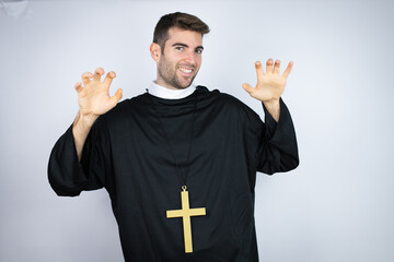 Young hispanic man wearing priest uniform standing over white background smiling funny doing claw...