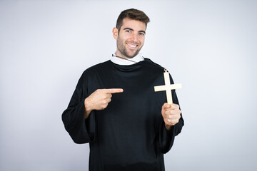 Young hispanic man wearing priest uniform standing over white background holding crucifix smiling...