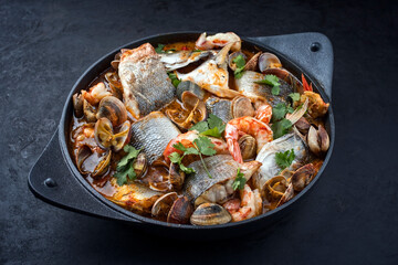Modern style traditional Spanish seafood zarzuela de pescado with fish, king prawns and venus clams served in clear sauce as close-up in design pot