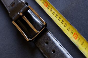 A black leather belt with holes and a metal buckle rests on a gray background next to a yellow tape...