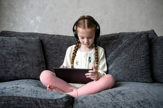 Beautiful cute you girl holding tablet computer sat on sofa watching screen wearing wireless technology headphones on cosy setee. Kid with pigtails smiling enjoying game in home lifestyle living room.